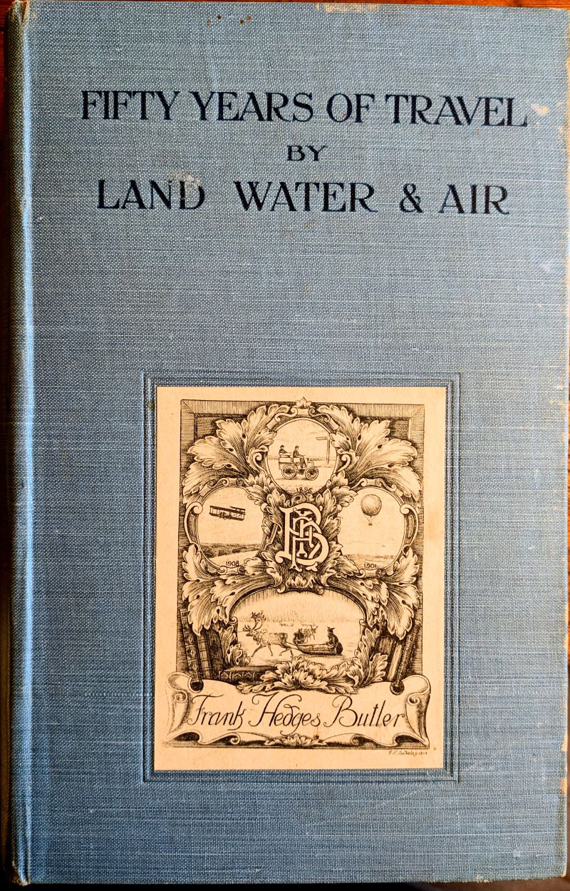 Cover of book showing title in black text bearing the author's bookplate with rich design of balloon, aeroplane, motor car and sledge pulled by Reindeer and his initials as a cipher.